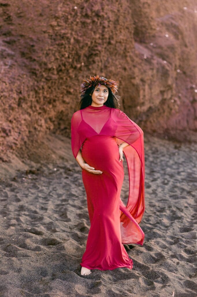 A woman in a flowing dress standing before the red cinder cone cliffs of a Maui black sand beach for her maternity photos