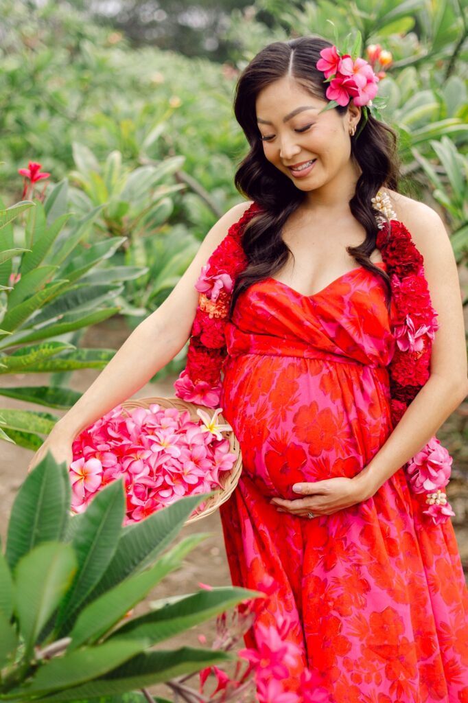 Editorial Maui babymoon maternity photos at a plumeria farm with a striking red and pink dress and floral lei