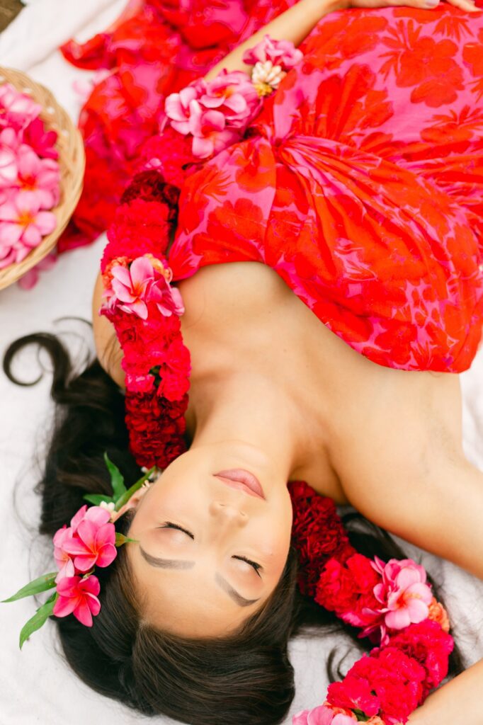 Expecting mother lying on a blanket surrounded by pink plumeria flowers during an editorial Maui babymoon session