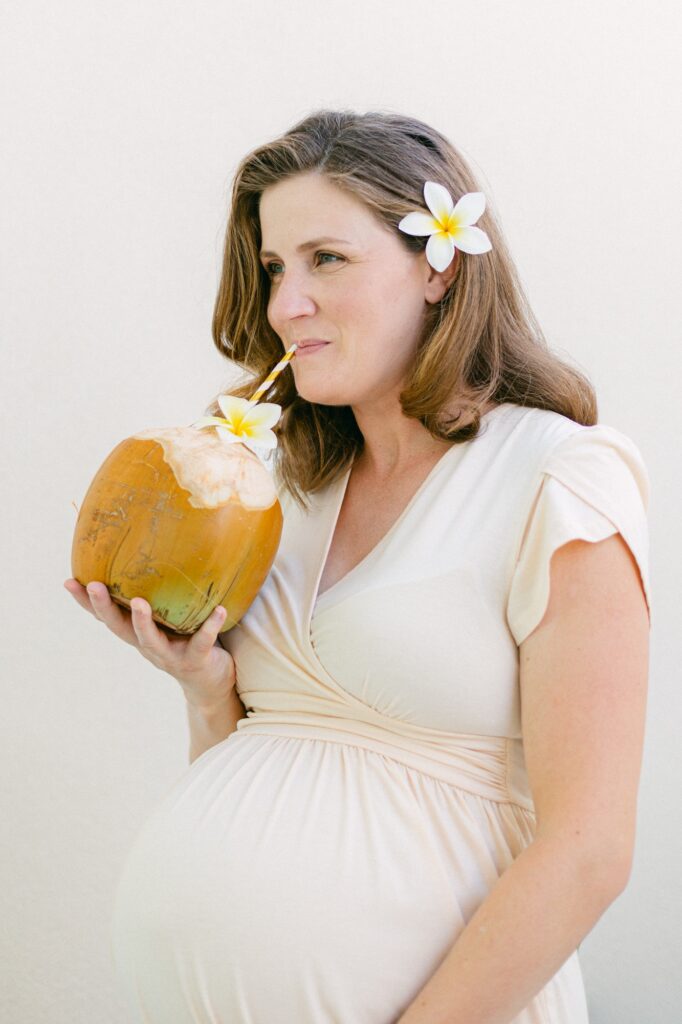 Expectant mother drinking fresh coconut juice on a Maui beach with a plumeria flower in her hair during a babymoon