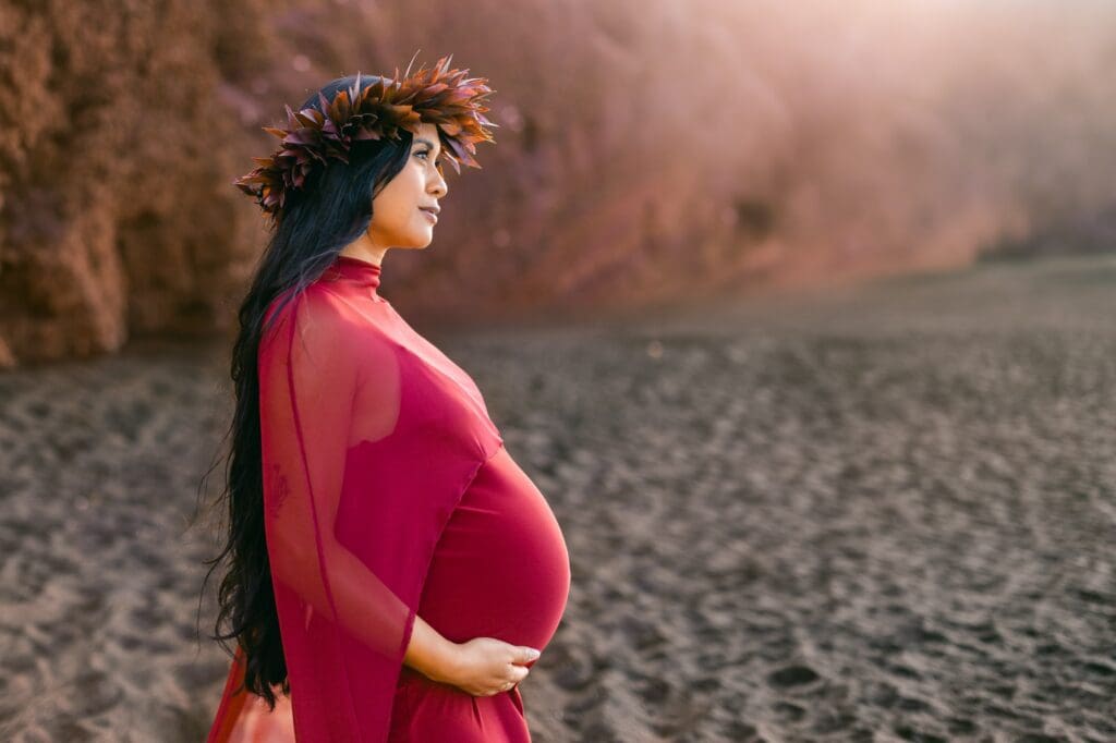 An expectant mother wearing a deep burgundy ti leaf haku lei on the volcanic black sand beach in Maui