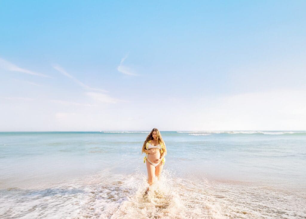 An expectant mother laughing and splashing in the shallow, calm water of Ukumehame Beach at sunset