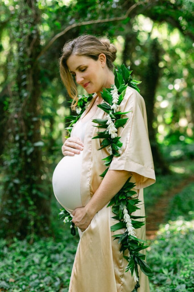 Expectant mother standing in the lush Honolua rainforest wearing a maile and tuberose lei