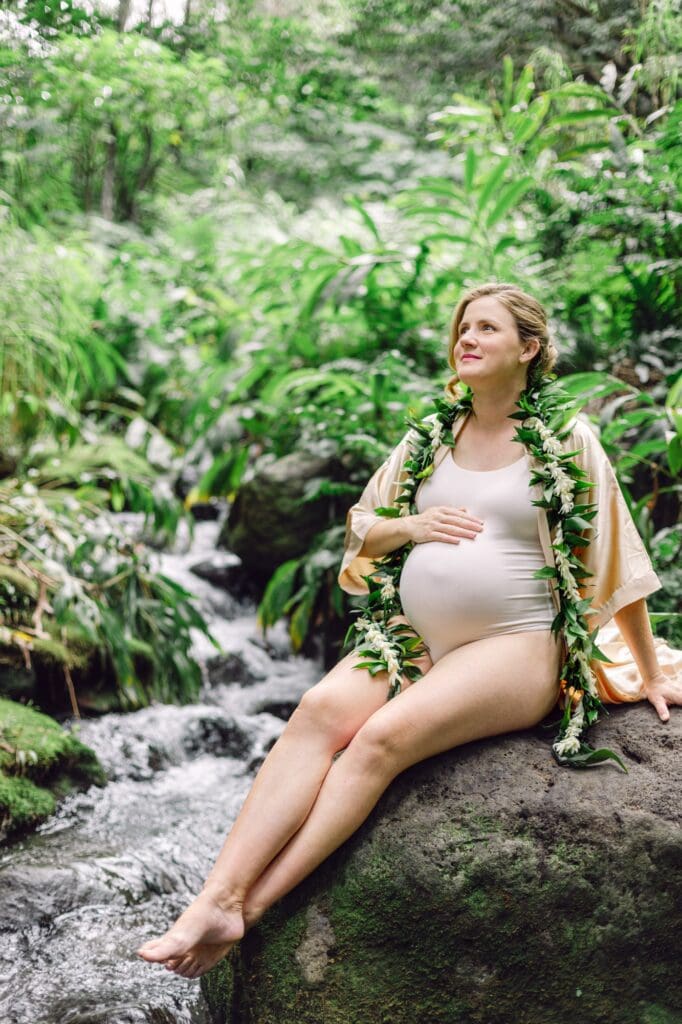 Pregnant woman seated by a forest stream in Iao Valley wearing a maile and tuberose lei