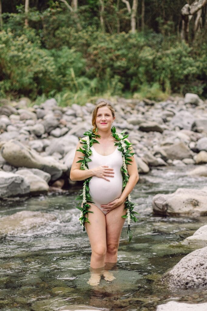 Maui maternity session in Iao Valley with expectant mother standing in a forest stream wearing a maile and tuberose lei