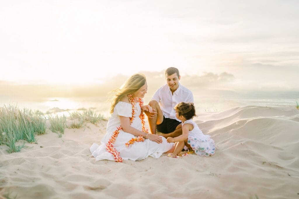 An expectant mother wearing a plumeria hapai lei sitting on the sweeping sand dunes at Ironwoods Beach