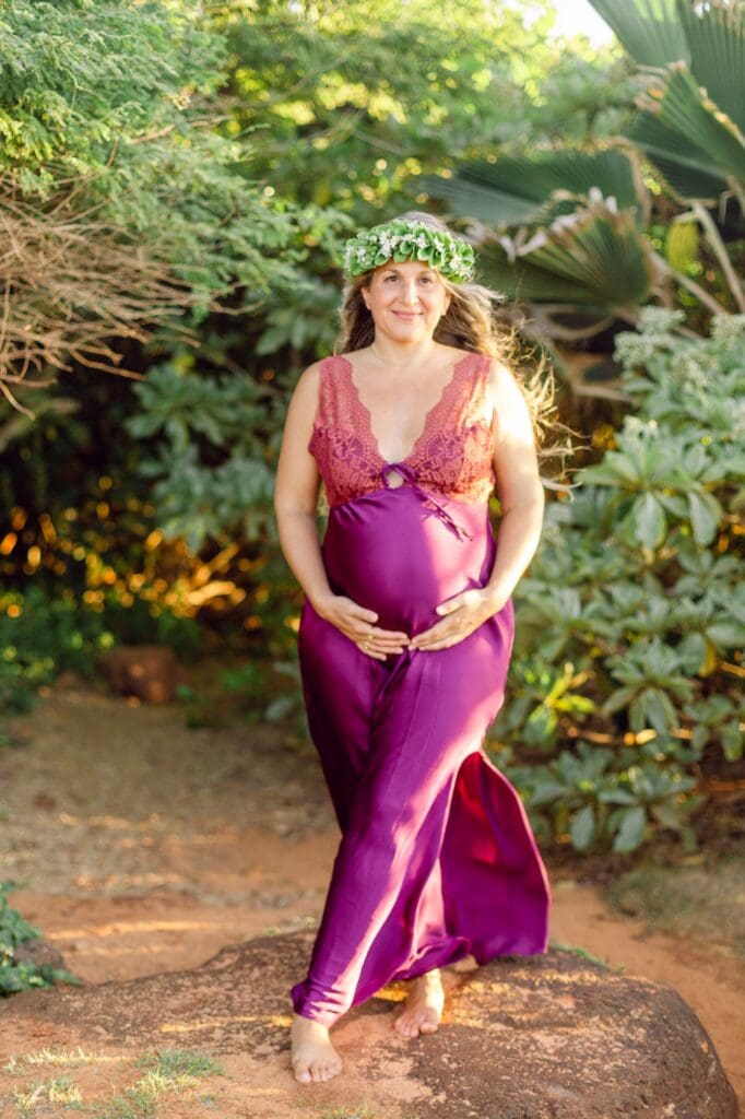 A babymoon portrait of an expectant mother framed by vibrant green naupaka shrubs and the red volcanic sand of Maui