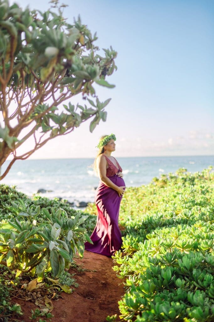 pregnant woman standing among naupaka on Maui red sand beach with ocean behind in wind