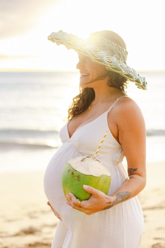 mother in papale hat with sun flare behind her holding coconut on Maui beach during maternity photo session