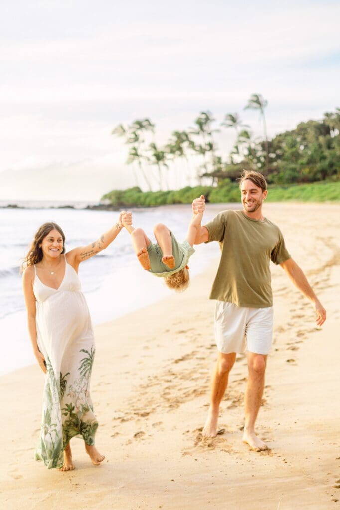 parents swinging toddler between them while walking on Maui beach during maternity photos