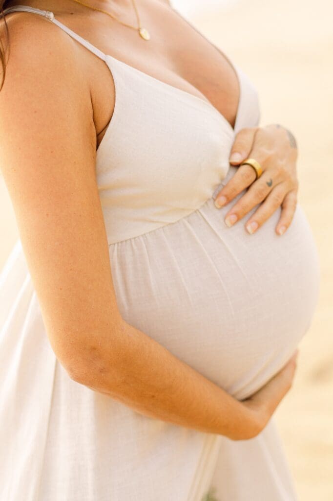 Detail shot of a white linen maternity dress and hands holding a baby bump, captured in the soft light of a Maui sunset