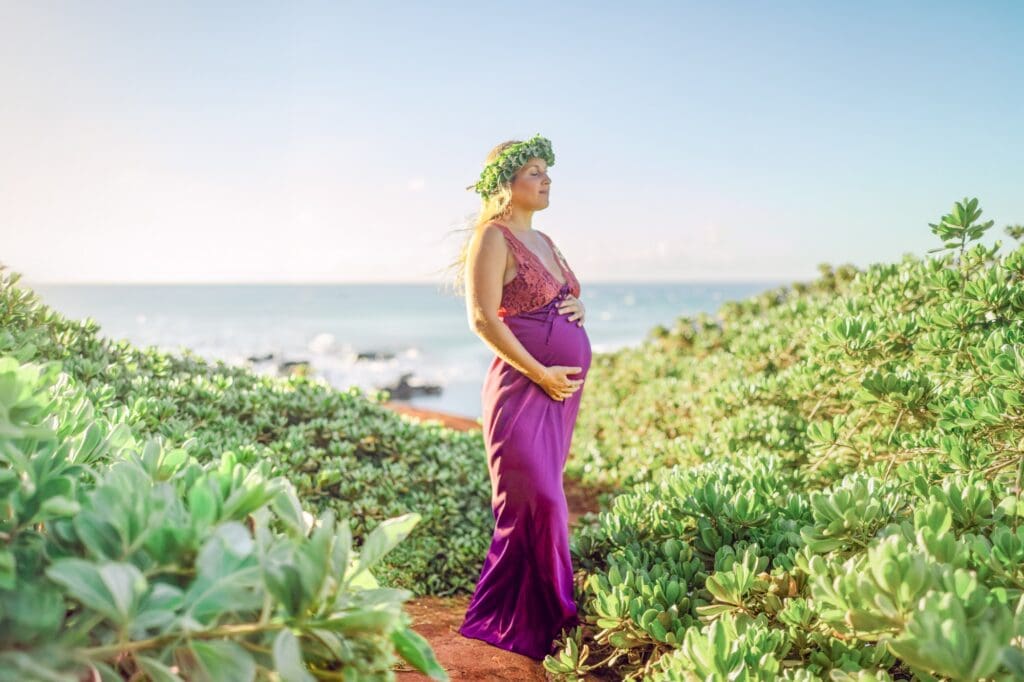 maternity photos on red sand beach Maui North Shore with ocean cliffs and green naupaka